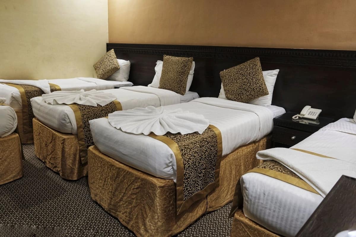 Une chambre d’hôtel avec quatre lits alignés, chacun étant doté de draps blancs et d’éléments décoratifs dorés. Une table de chevet avec un téléphone et une lampe se trouve sur le côté droit.