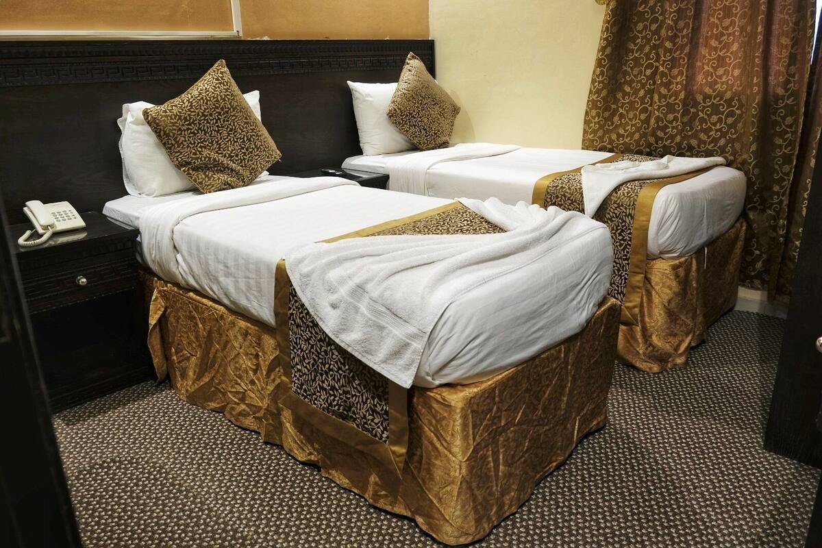 Une chambre d’hôtel avec deux lits, chacun recouvert d’une courtepointe blanche agrémentée de motifs dorés, un téléphone sur une table de chevet et des rideaux à motifs.