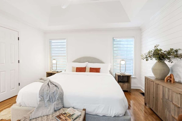 A bedroom with a large bed, two bedside tables, a nightstand, a wooden dresser, and a window with blinds.