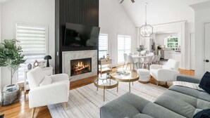 A modern living room with a fireplace, white furniture, and a dining area visible through an open door. - Luxury Retreat & Heated Pool Downtown Simpsonville (Simpsonville)