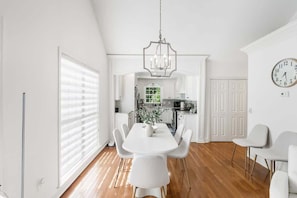 Une cuisine moderne avec des meubles blancs, un coin repas doté d’une table et de chaises blanches, et une grande fenêtre avec des stores.