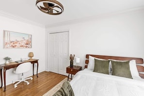 A bedroom with a wooden desk, a bed with white bedding, a wooden headboard, a white door, and a ceiling light fixture.