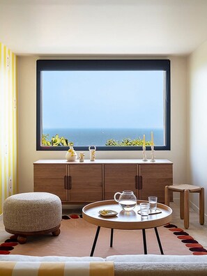 A room with a large window overlooking the sea, a round wooden table with a pitcher and glasses, a small stool, and a cabinet with decorative items. - Villa with a 180° ocean view, renovated in 2024—a little paradise in Rayol Canadel (Rayol-Canadel-sur-Mer)