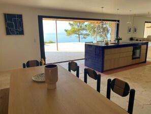 A modern kitchen with a blue tiled island, a dining area with a wooden table and chairs, and a large glass door leading to an outdoor terrace with a view of the sea.