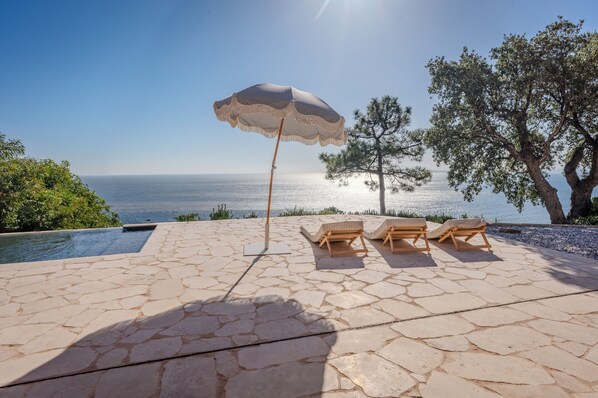 A stone patio with an umbrella and lounge chairs overlooking a pool and the sea. - 180° sea view villa, renovated in 2024, a little paradise in Rayol Canadel (Rayol-Canadel-sur-Mer)