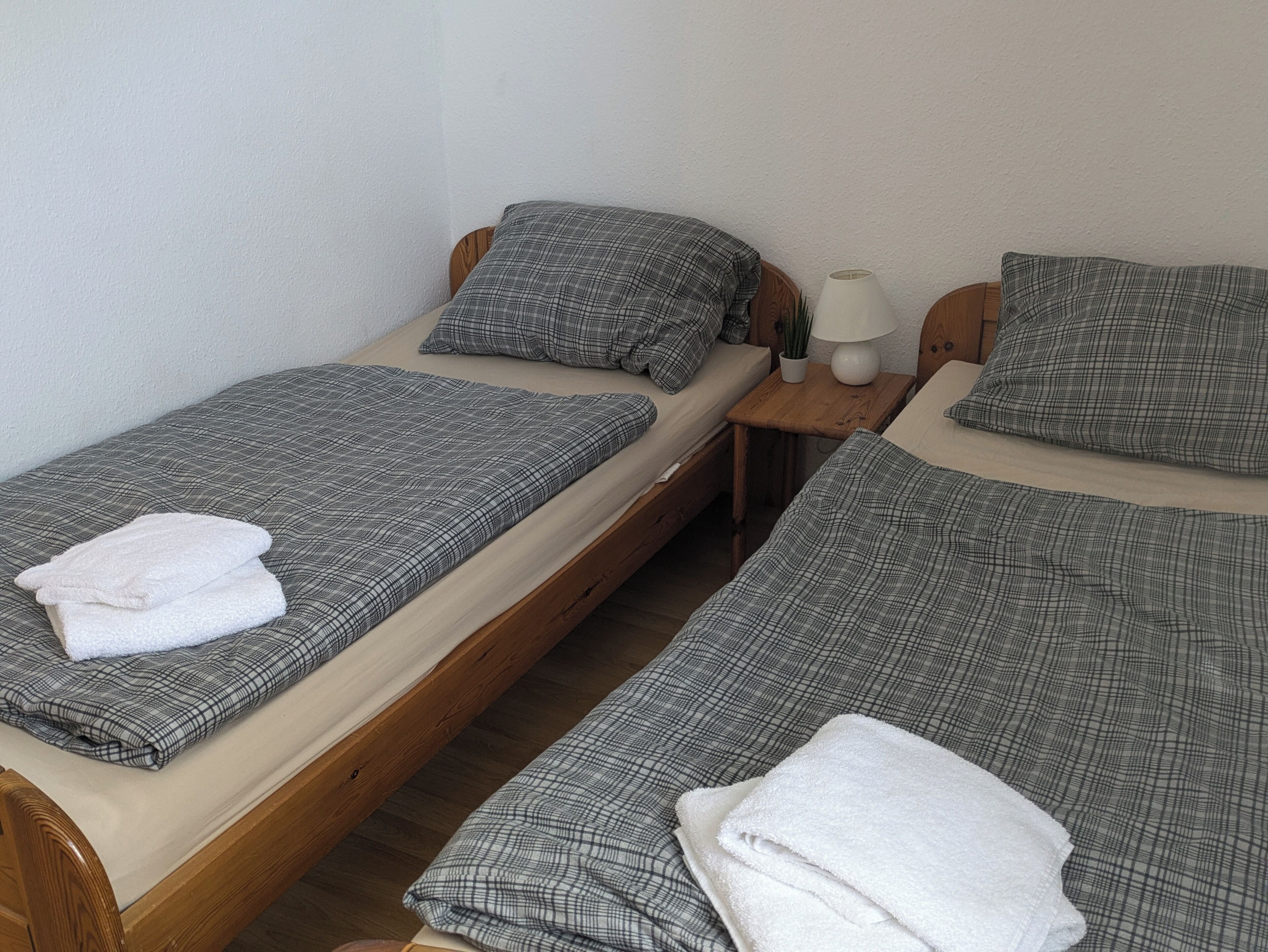 Dos camas individuales con ropa de cama de cuadros grises, cabeceras de madera y lámparas blancas sobre mesitas de noche de madera.