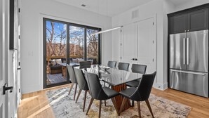 Una cocina moderna con un refrigerador de acero inoxidable grande, una zona de comedor con una mesa de vidrio y sillas negras, y vistas al exterior a través de la puerta de vidrio.