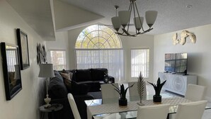 A modern living room with a glass dining table, white chairs, a black sofa, and a TV.