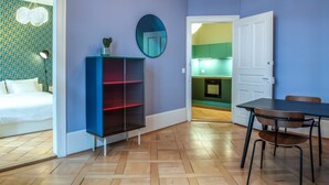 A room with a bed, a blue and red shelving unit, a dining table with chairs, and a kitchen visible through an open door. - ReLiving – Your Group’s Perfect Getaway (Luzern)
