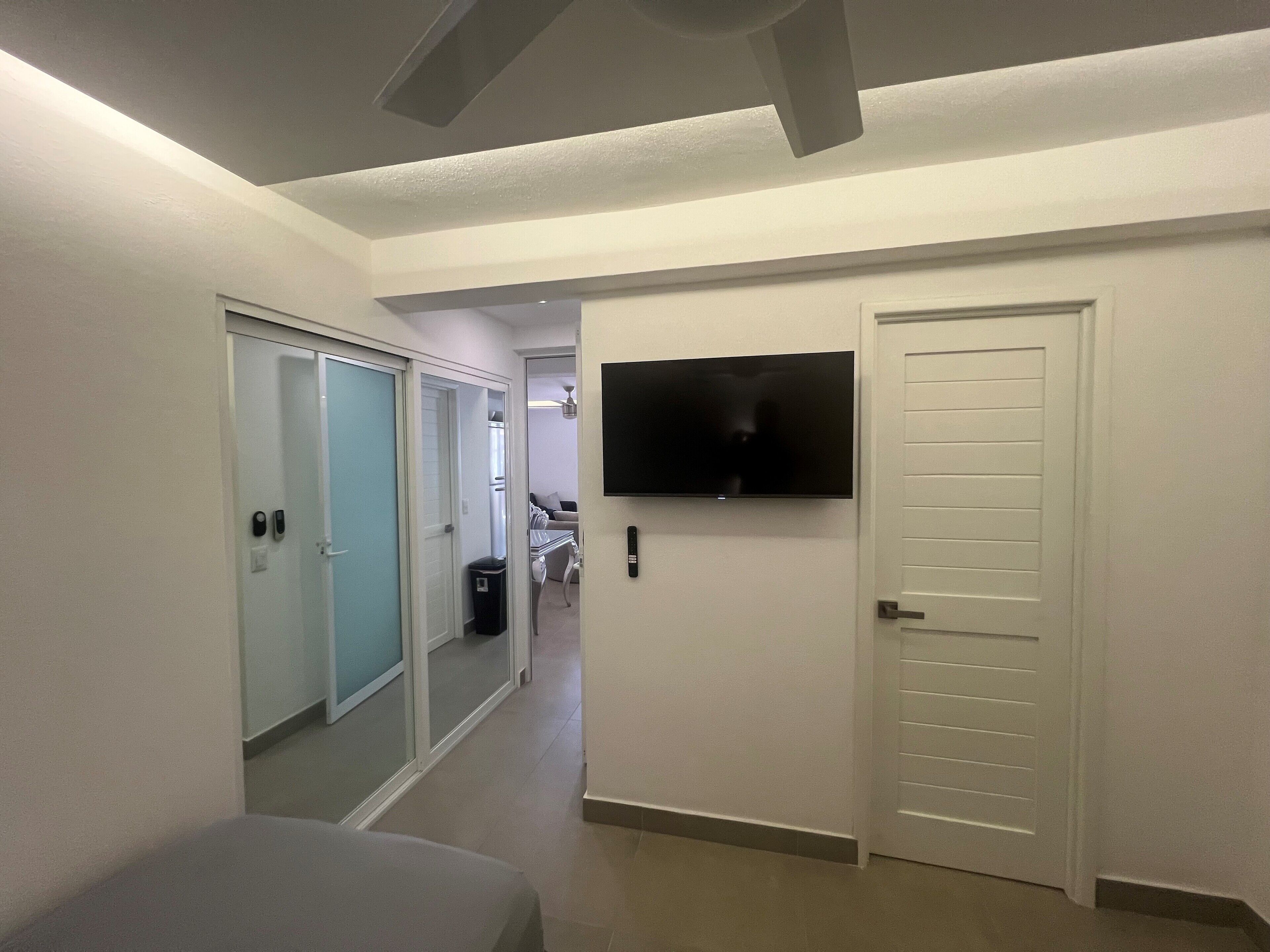 Ein Hotelzimmer mit einem Bett, einem an der Wand befestigten Fernseher, einer Tür und einem Flur, der zu einem anderen Zimmer führt.