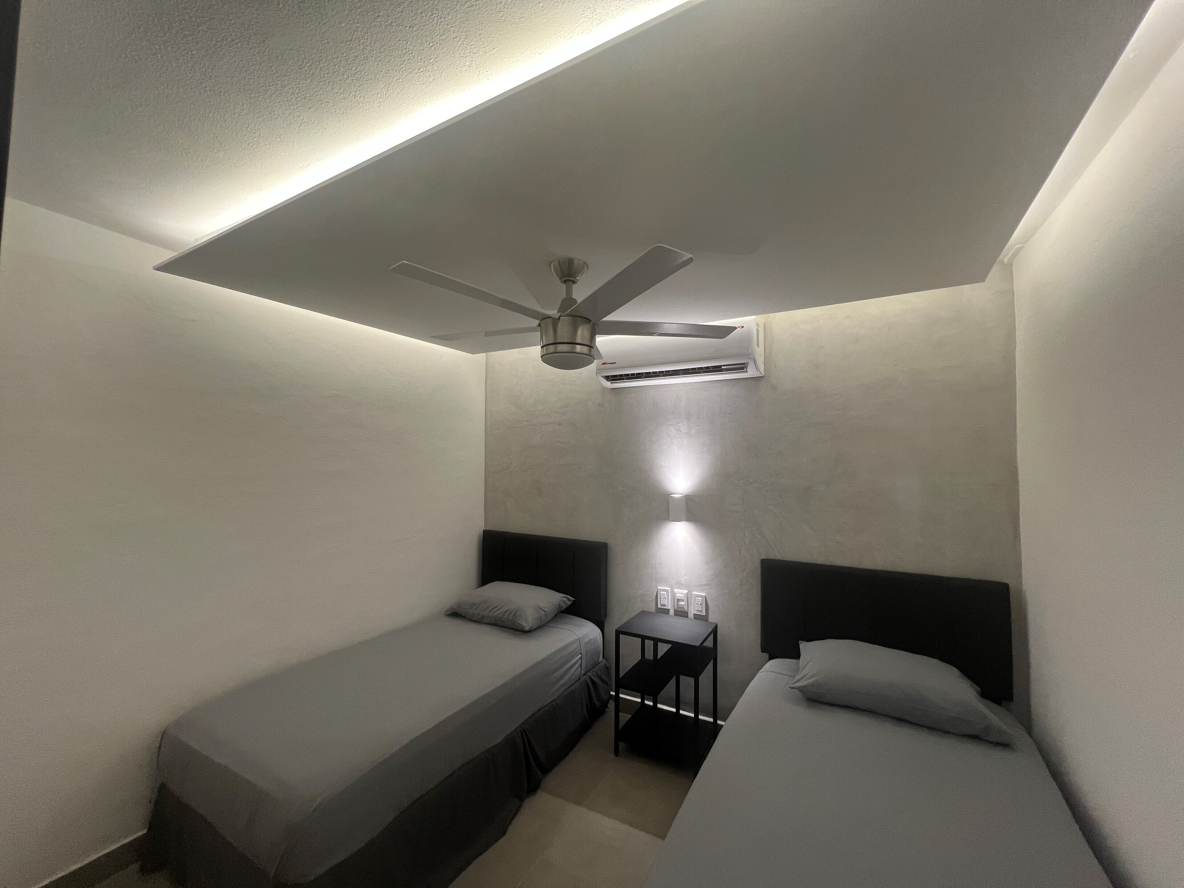 Quarto pequeno e pouco iluminado, com duas camas de solteiro, um ventilador de teto e um aparelho de ar condicionado.