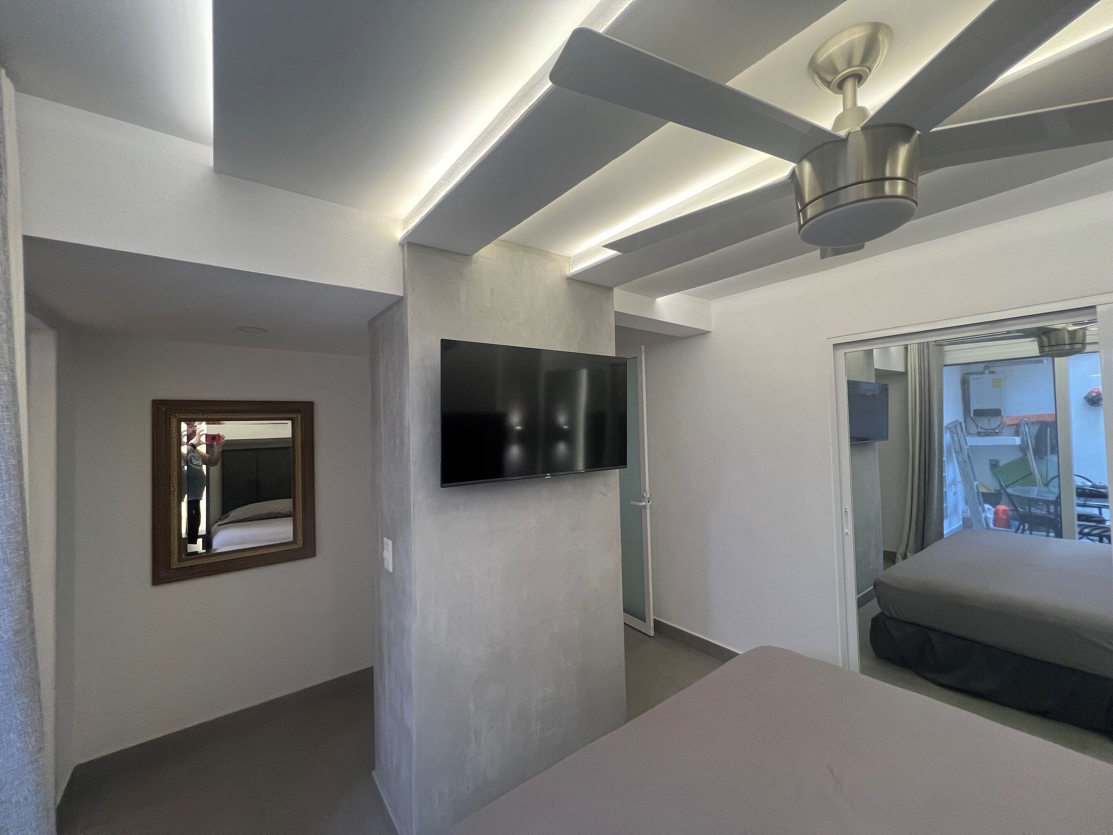 A modern hotel room with a flat-screen TV mounted on the wall, a ceiling fan, and a bed with a mattress and pillows.