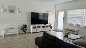Ein modernes Wohnzimmer mit einem großen Flachbildfernseher, einem dunklen Sofa, einem kleinen weißen Tisch und einem Deckenventilator.