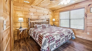 Un dormitorio de cabaña de madera con una cama, dos mesitas de noche, un ventilador de techo y una ventana con persianas.