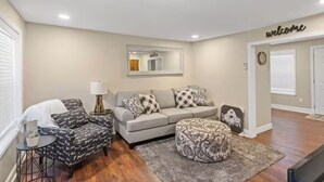 A living room with a grey sofa, patterned ottoman, and a small side table with a lamp. - Townhouse near WKU & Mammoth Cave (Bowling Green)