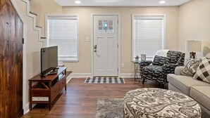 A living room with a television on a wooden stand, a sofa with patterned cushions, and a patterned ottoman. - Townhouse near WKU & Mammoth Cave (Bowling Green)