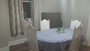 En matplats med ett runt bord täckt av en rutig duk, omgivet av fyra vita stolar med träramar. Det finns ett fönster med gardiner, ett träskåp och en liten krukväxt på bordet.