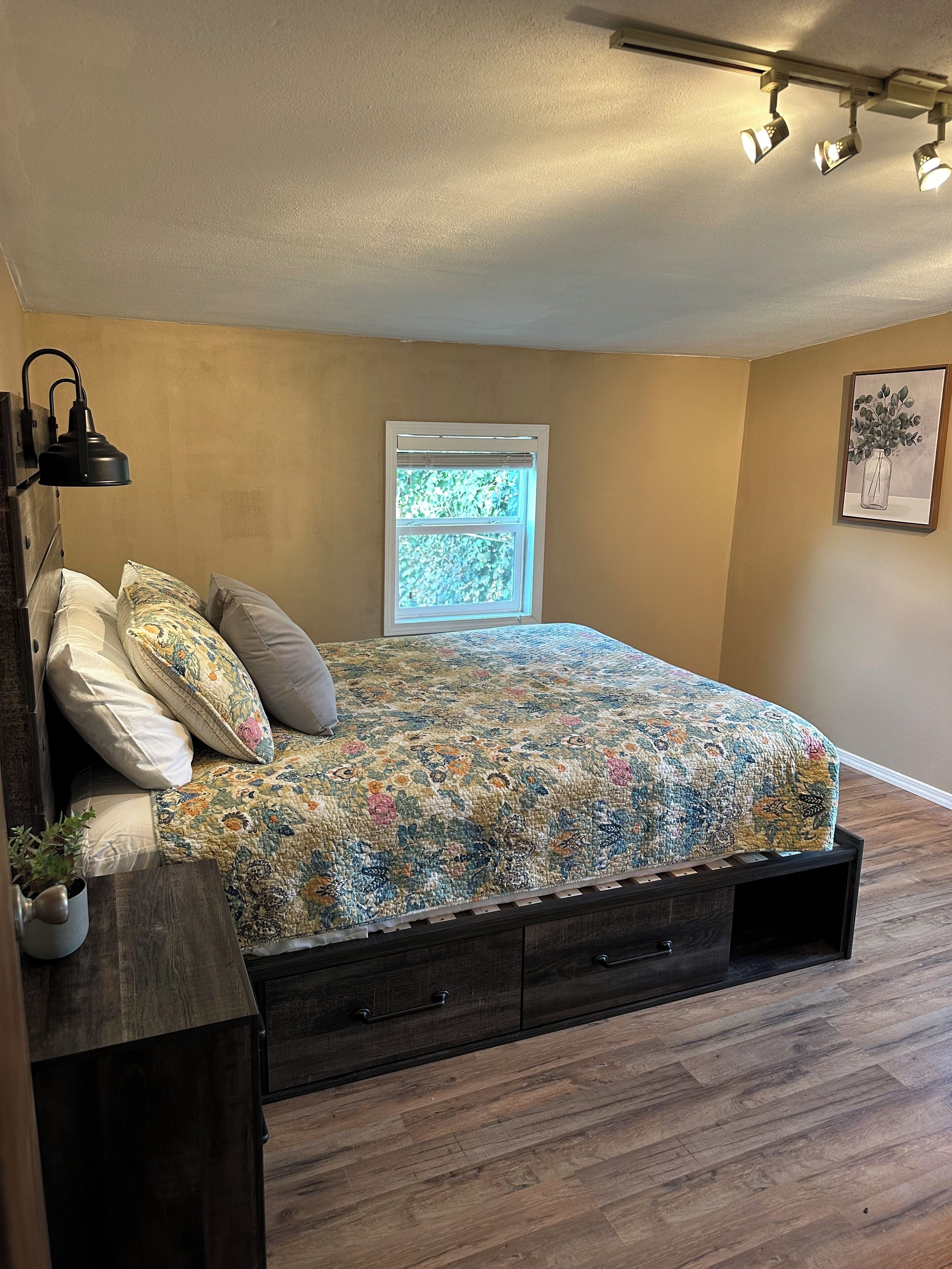 A bedroom with a bed, a window, a wall-mounted lamp, and a framed picture.