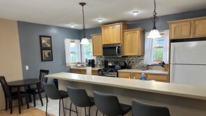 A modern kitchen with wooden cabinets, a central island with bar stools, a microwave, and a refrigerator. - Custer Cottage / Fenced Yard (Billings)