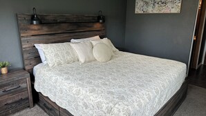 A bedroom with a wooden headboard, a large bed with patterned bedding, a nightstand with drawers, a small plant, and a floral painting on the wall. - Custer Cottage / Fenced Yard (Billings)