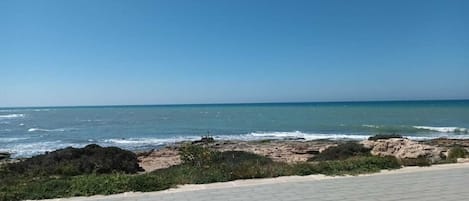 Vista costera con un paseo pavimentado que conduce a la playa, terreno rocoso y el océano.