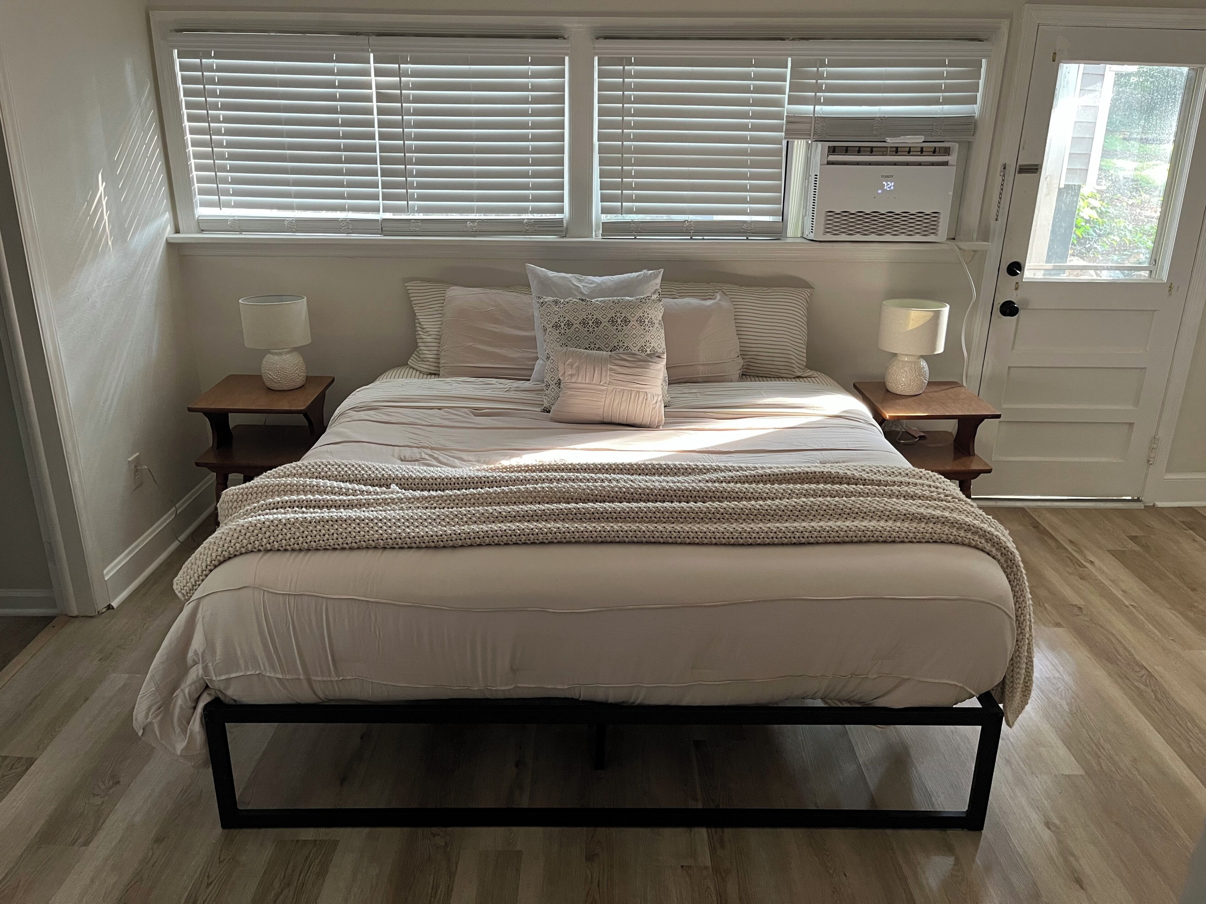 Ein Schlafzimmer mit einem Bett, zwei Nachttischen, einem Fenster mit Jalousien, einer Klimaanlage und einer Tür.