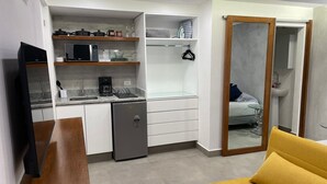 A compact living space with a kitchenette, a small refrigerator, a microwave, and a coffee maker. There is a bed with a glass side table and a yellow armchair.