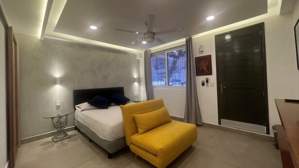 A modern bedroom with a bed, a yellow armchair, a ceiling fan, and a window with curtains.