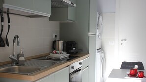 Cocina compacta con lavabo, fogón y microondas. Hay una mesa con una bandeja de vasos rojos y una silla.