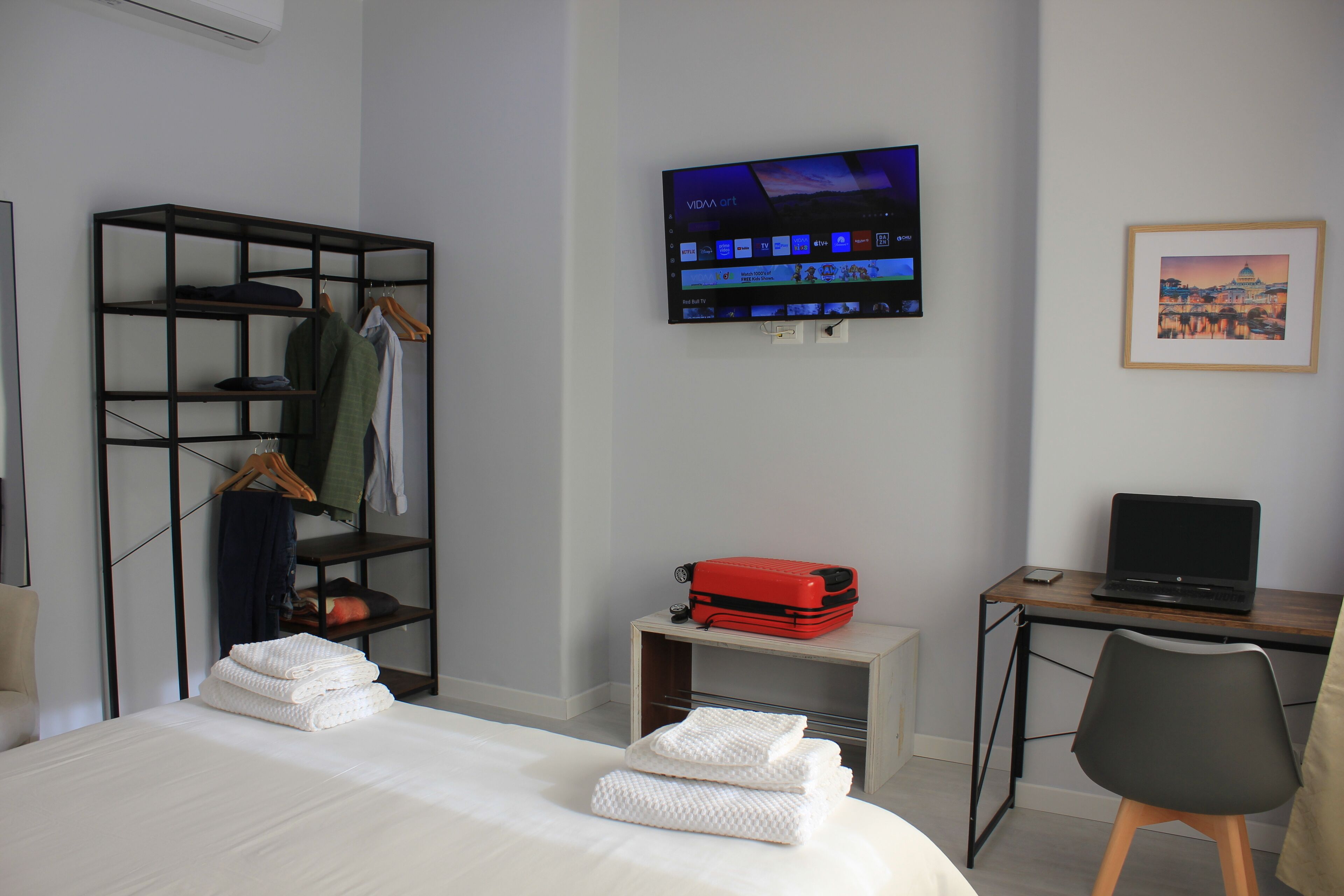 Ein Hotelzimmer mit Bett, Schreibtisch mit Laptop, Wandfernseher, Kleiderschrank und einem gerahmten Bild.