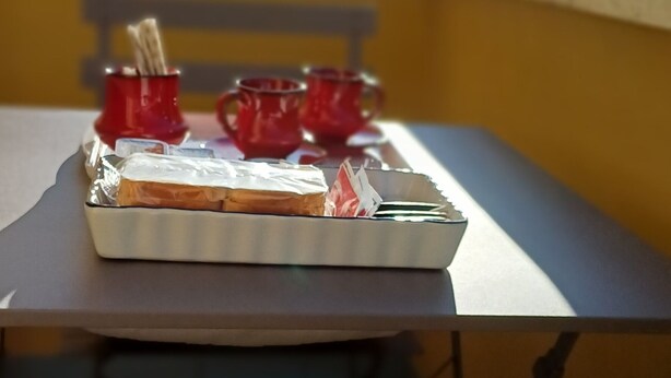 Una mesa con una caja de regalo envuelta, vasos rojos, una ventana con vistas a un edificio y una silla con mantel navideño.