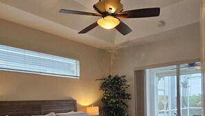 Ein Schlafzimmer mit einem großen Bett, einem Deckenventilator, einer Topfpflanze und Blick ins Freie durch eine Schiebetür.