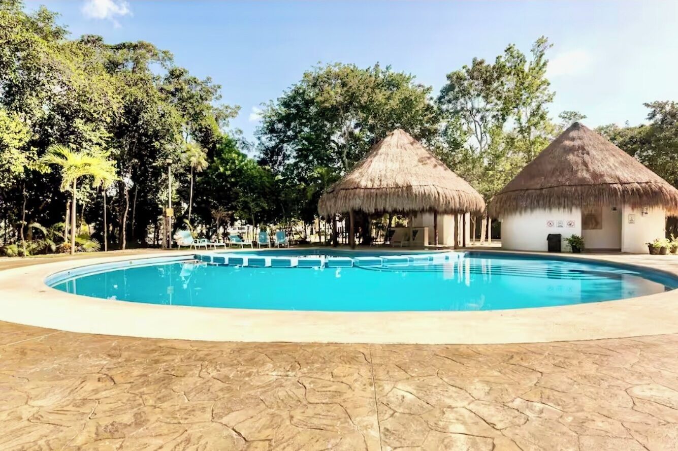 A swimming pool with two thatched-roof huts, surrounded by trees.