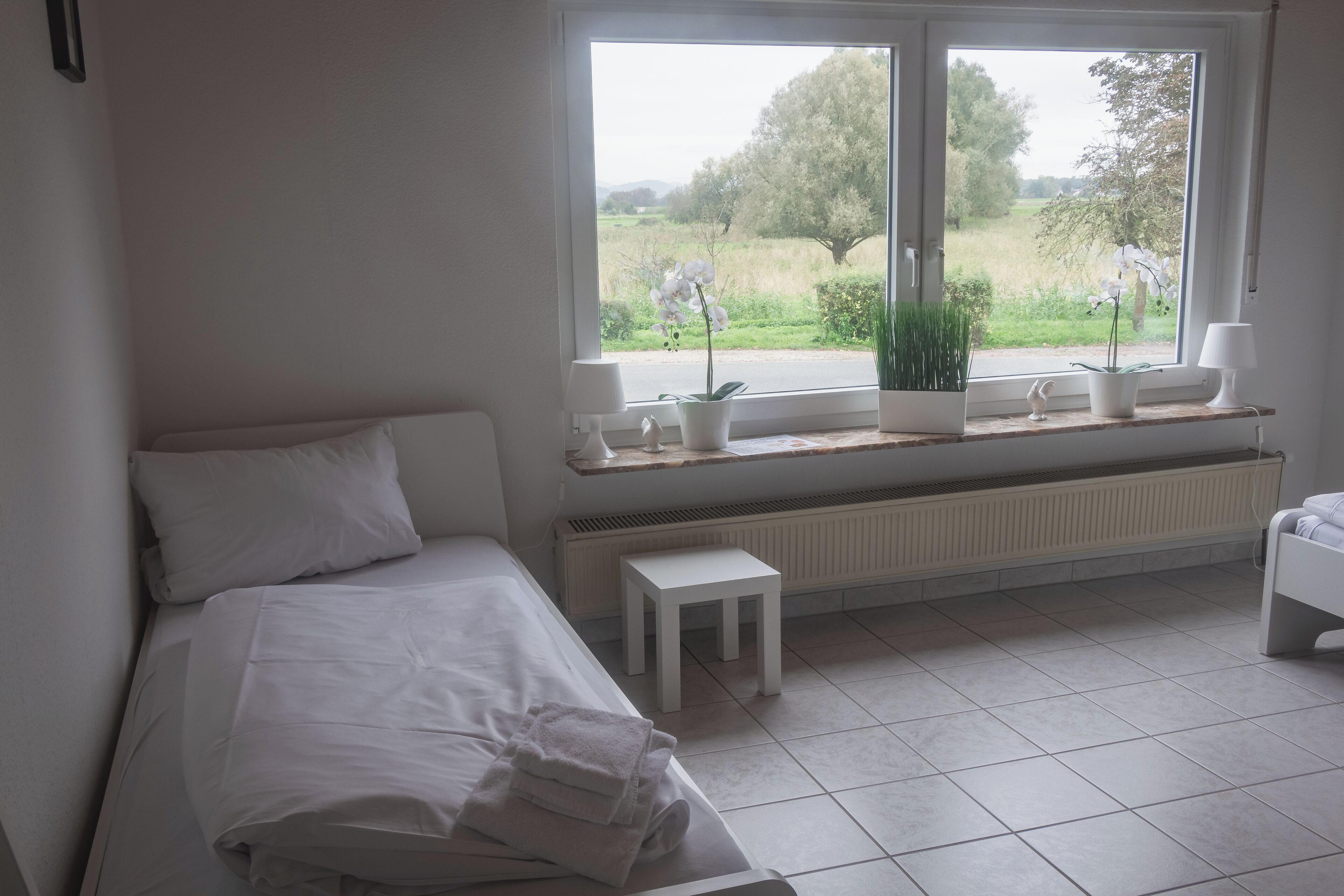 A bedroom with a bed, a window with a view of greenery, a radiator, and a small table.