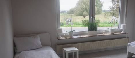 A bedroom with a bed, a window with a view of greenery, a radiator, and a small table.