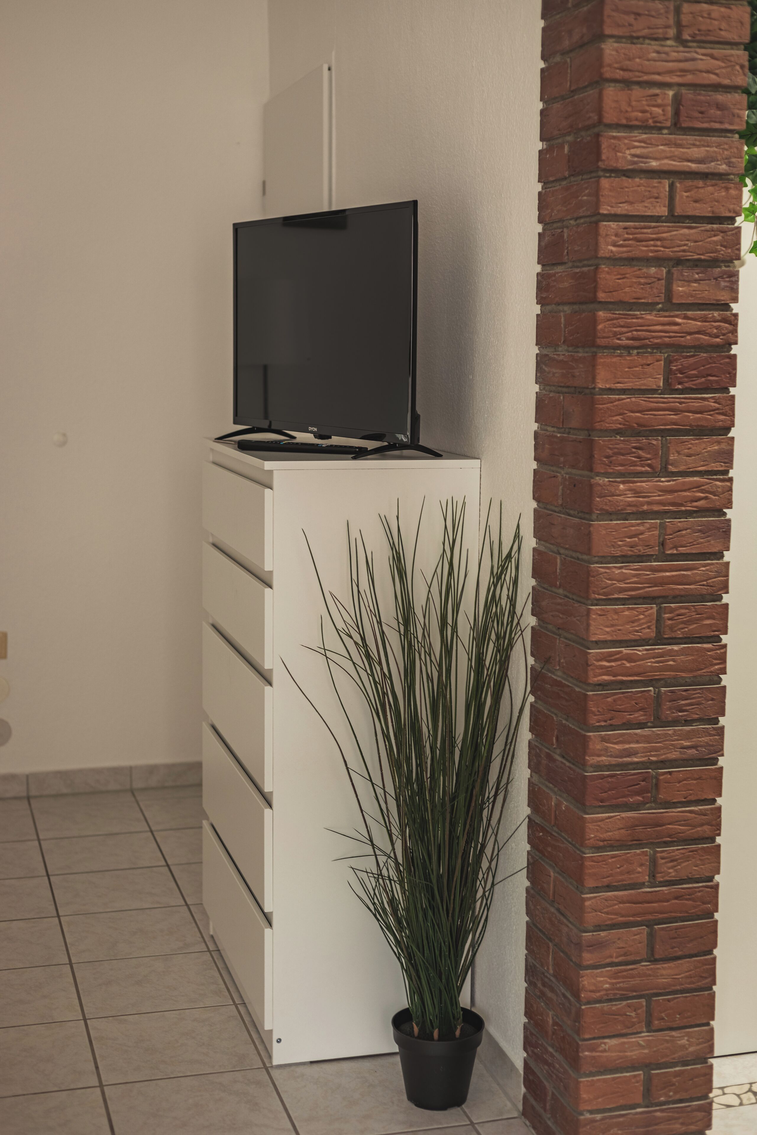 A white entertainment unit with a flat-screen TV on top, a potted plant beside it, and a brick column to the right.