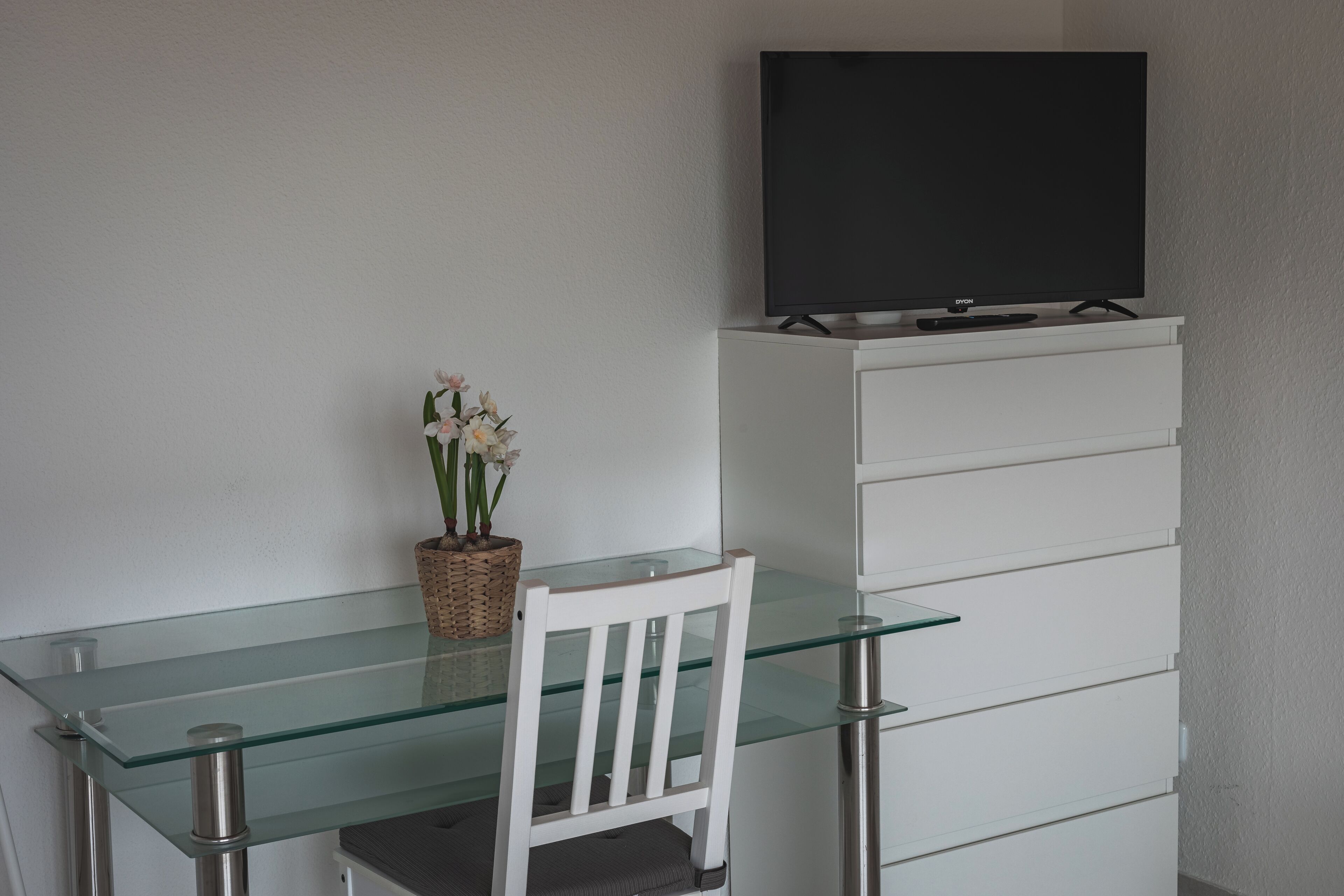 Une chambre moderne avec un bureau en verre, une chaise blanche, un téléviseur posé sur une commode blanche et un panier en osier rempli de fleurs.