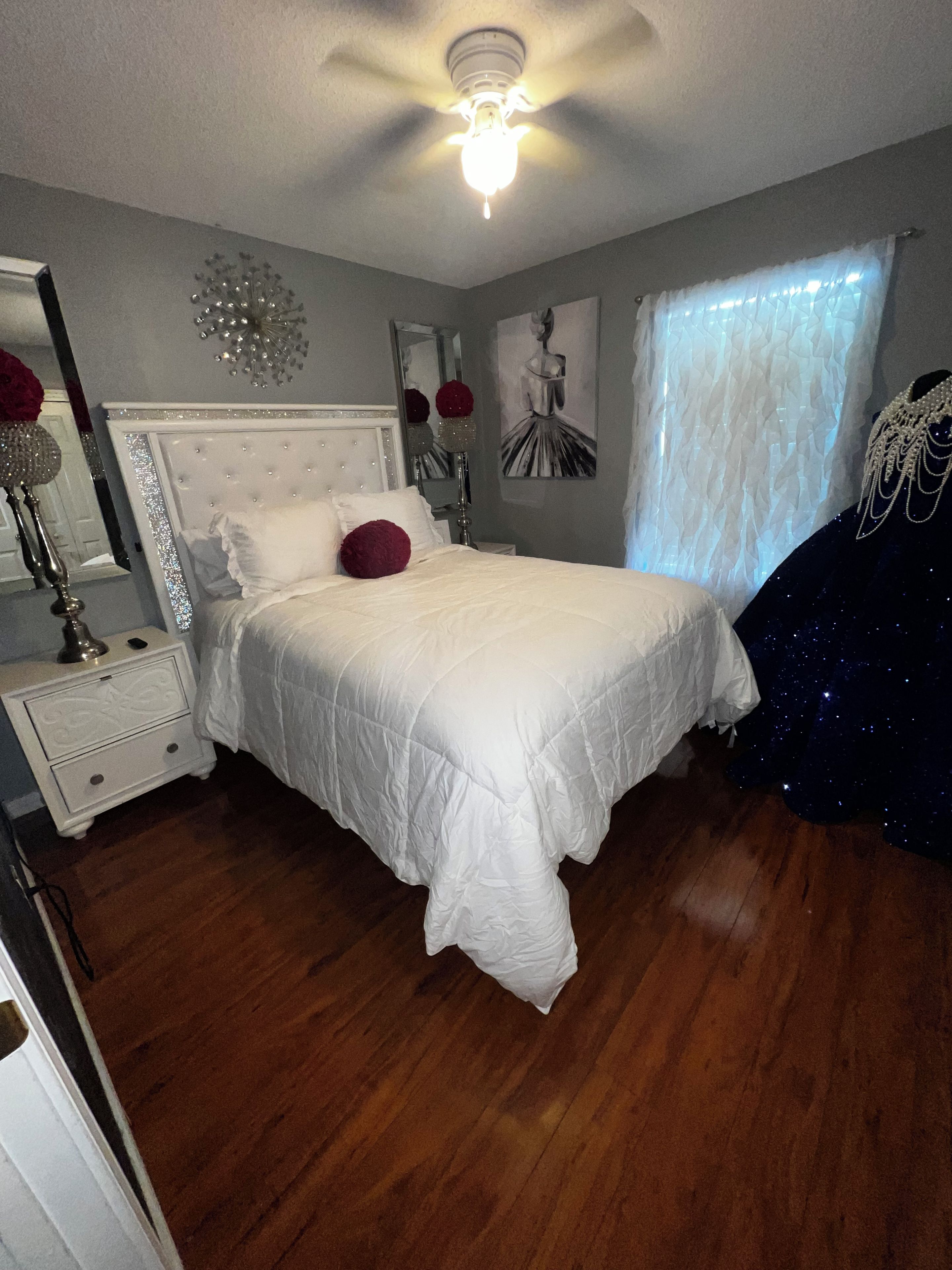 A bedroom with a bed, a nightstand, a mirror, a ceiling fan, and a window with sheer curtains.