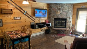 A cozy living room with a stone fireplace, wooden walls, a dining area with a table and chairs, and a television mounted on the wall. - Cozy Condo, 3 beds, 2 baths, kitchen, near RMNP, Hiking, Golf, Skiing and more! (Granby)