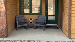 A porch with two wicker chairs and a small table, a bottle of wine, and two green-framed windows. - Cozy Condo, 3 beds, 2 baths, kitchen, near RMNP, Hiking, Golf, Skiing and more! (Granby)
