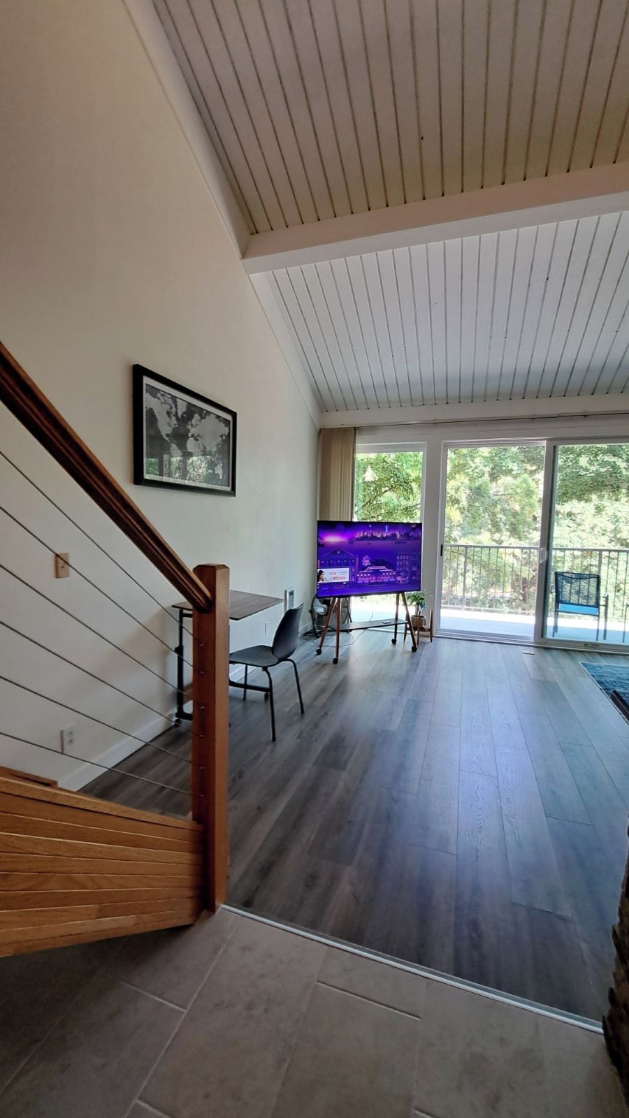 A spacious room with a wooden staircase, a large flat-screen TV, and a balcony with a view of trees.