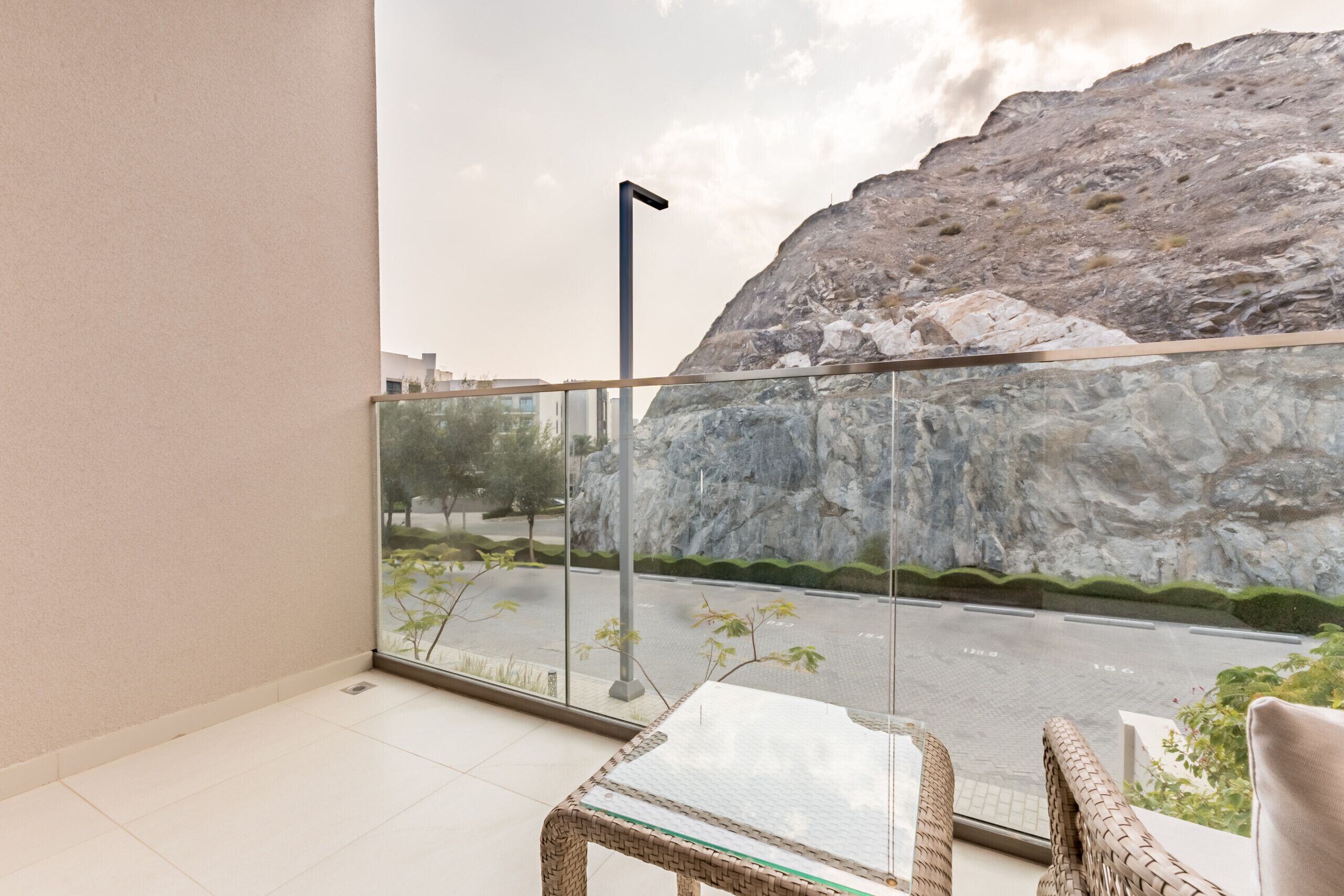 A balcony with a glass railing, a wicker chair, and a view of a rocky landscape and road.