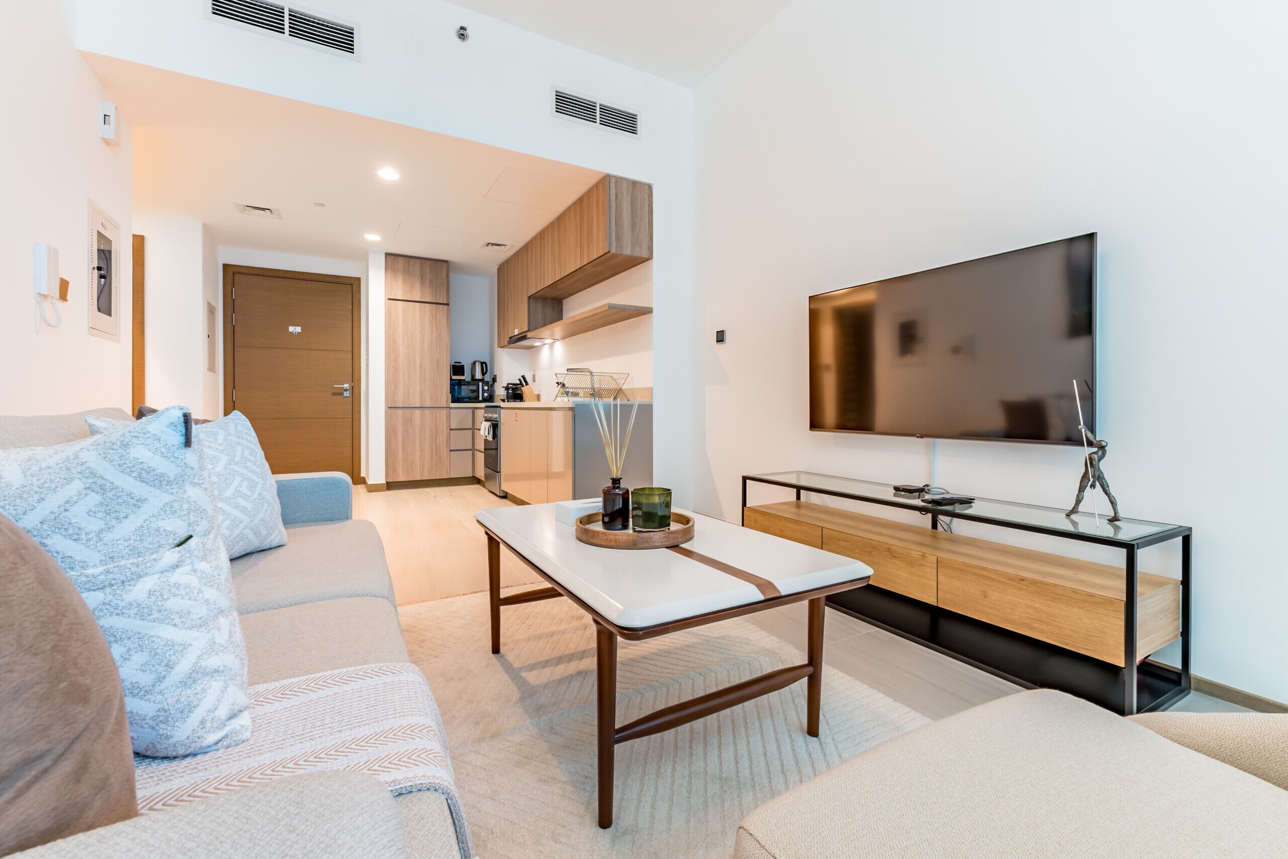 A modern living room with a sofa, coffee table, and TV. There is a kitchen in the background with wooden cabinets and a dining area.