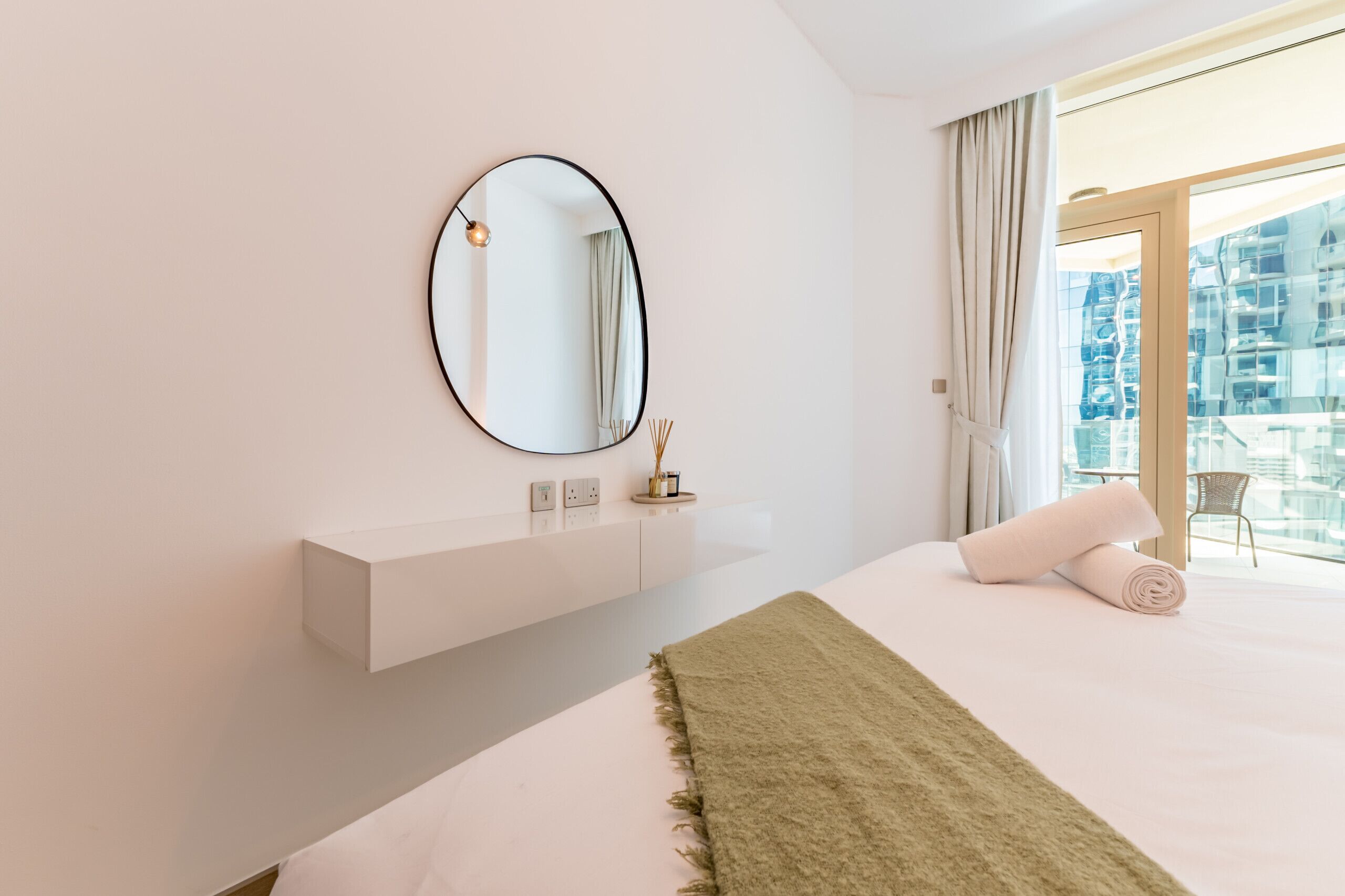 Une chambre d’hôtel moderne avec un grand lit, un miroir rond, une table de chevet blanche et une vue sur la ville grâce à de grandes fenêtres.