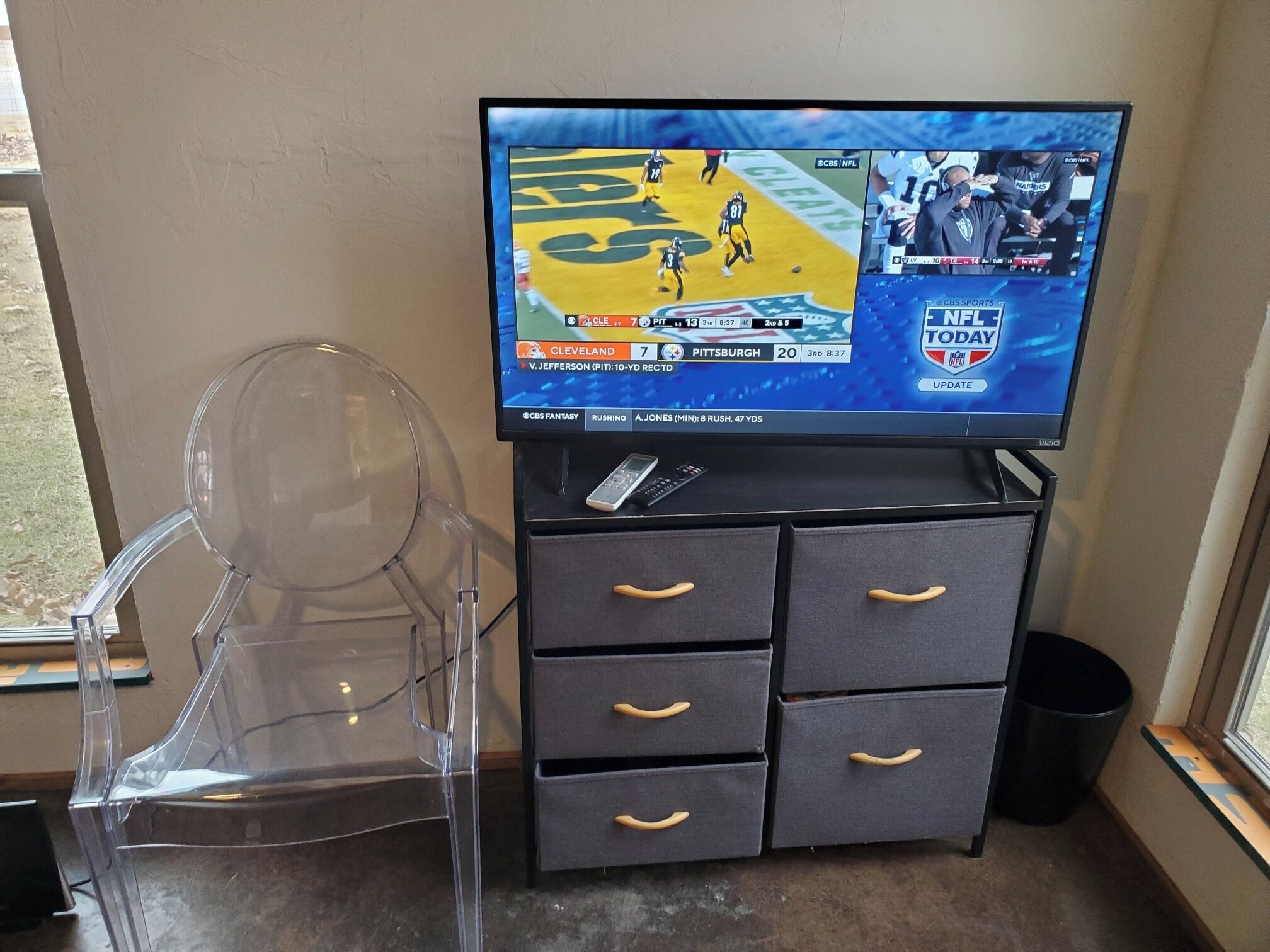 Un téléviseur diffusant un match de football, une chaise en plastique transparent et un petit meuble noir avec des tiroirs.