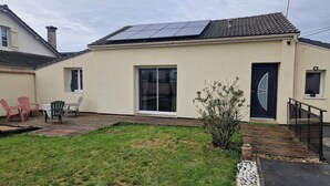 Una casa de una planta con tejado de tejas, un amplio ventanal corredero y un pequeño jardín con terraza de madera y una planta en maceta.