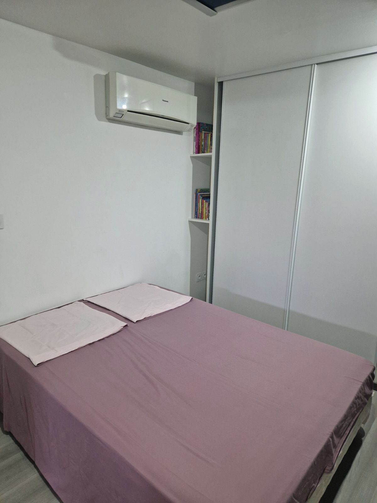 A bedroom with a bed, an air conditioner, a wardrobe, and a bookshelf with books.