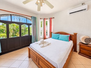 A bedroom with a bed, a ceiling fan, an air conditioning unit, and a view of greenery through the window. - 7BR · 2 Homes · Sleeps 16 · Pool ·  Walk to Jacó Beach (Jaco)