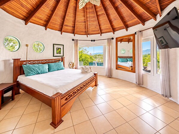 A bedroom with a wooden bed, a nightstand, a large window with a view, and a ceiling with wooden beams.