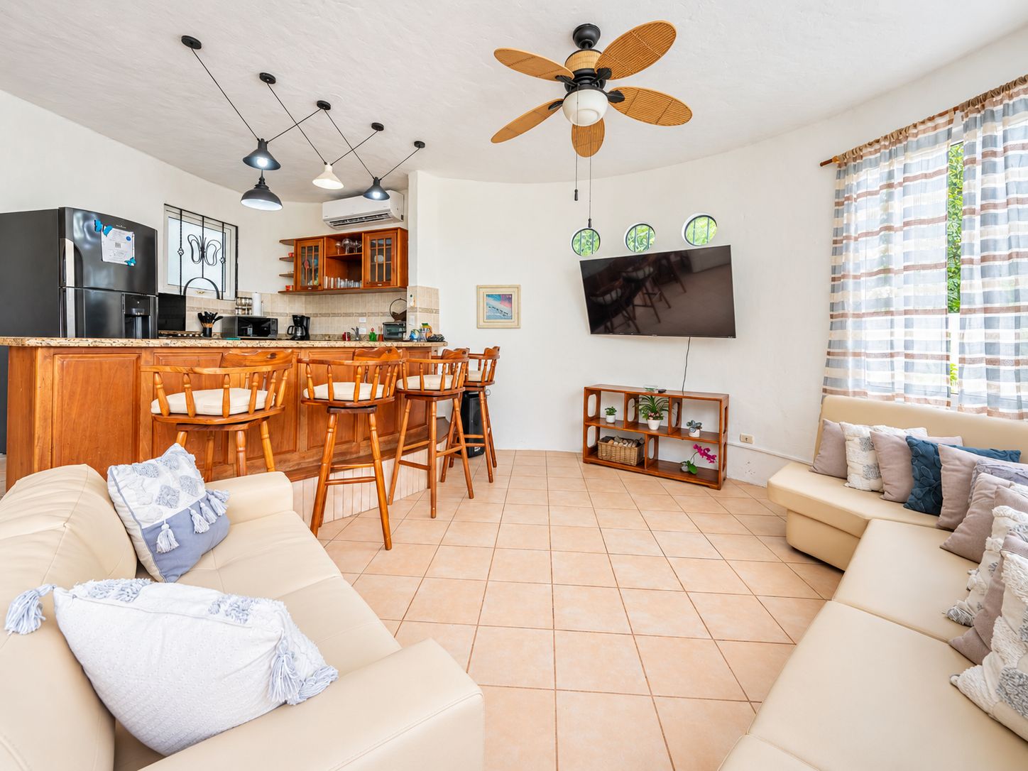 A modern living room with a sofa, a dining area with chairs, a kitchen with a refrigerator, and a ceiling fan.